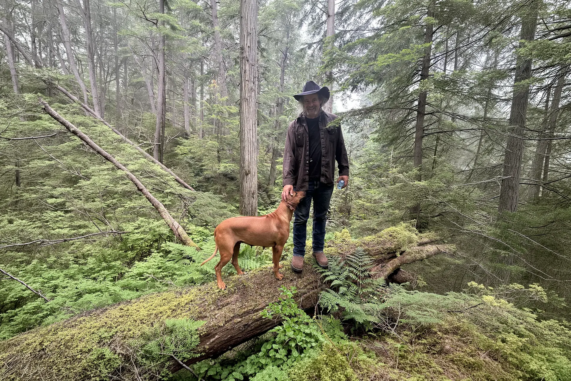 Founder Lawrence Dunkley standing on a mossy log in an old-growth forest with Jakira, his dog